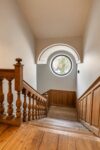 Escalier historique en bois du Manoir avec rambarde sculptée et fenêtre ronde.